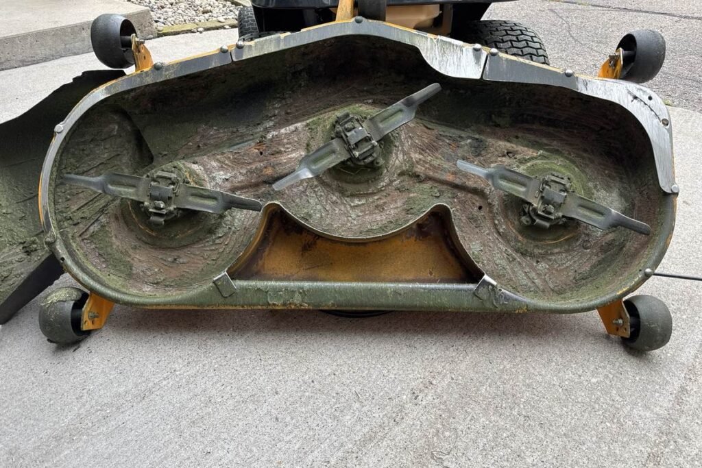 Underside of a three-blade riding mower deck flipped on a driveway, showing blades, anti-scalp rollers, and caked grass buildup
