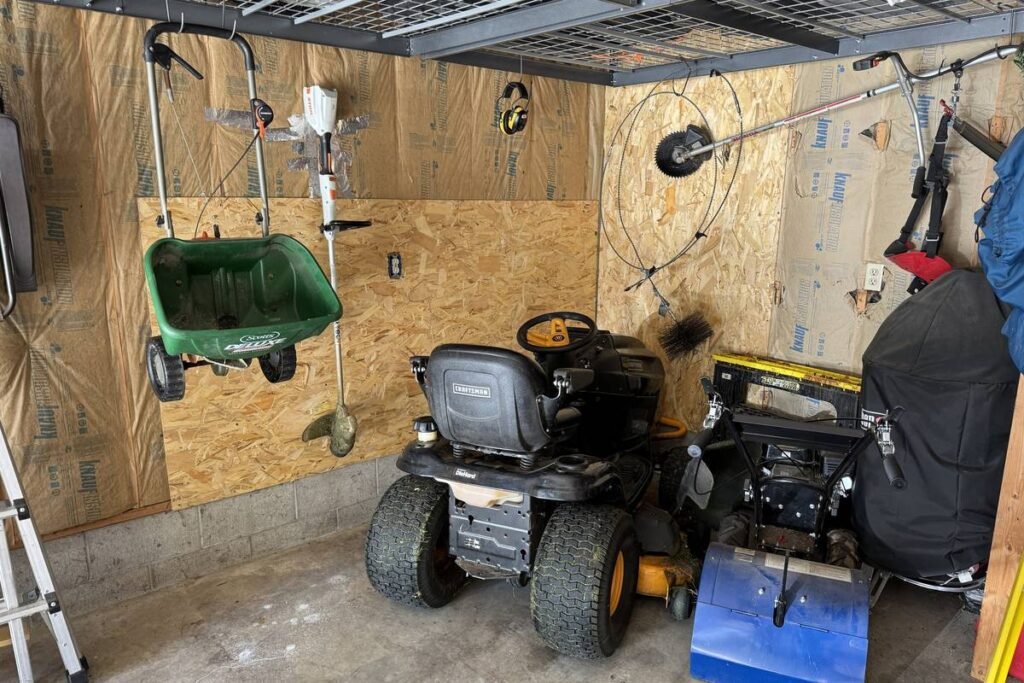 Lawn equipment stored in a garage — rider mower, broadcast spreader, string trimmer and rototiller